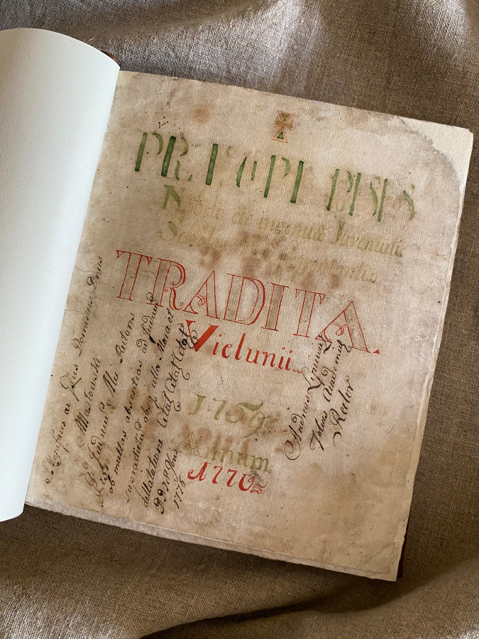 Otwarta księga Principia poesis. Na stronie tytułowej łaciński tekst, litery w kolorach: zielonym, czerwonym i czarnym. W tle drapowane płótno. 